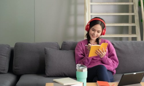 teenager wearing headphones and writing while sitting on sofa