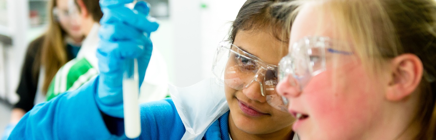 Two girls wearing goggles and lab coats holding up a vial.
