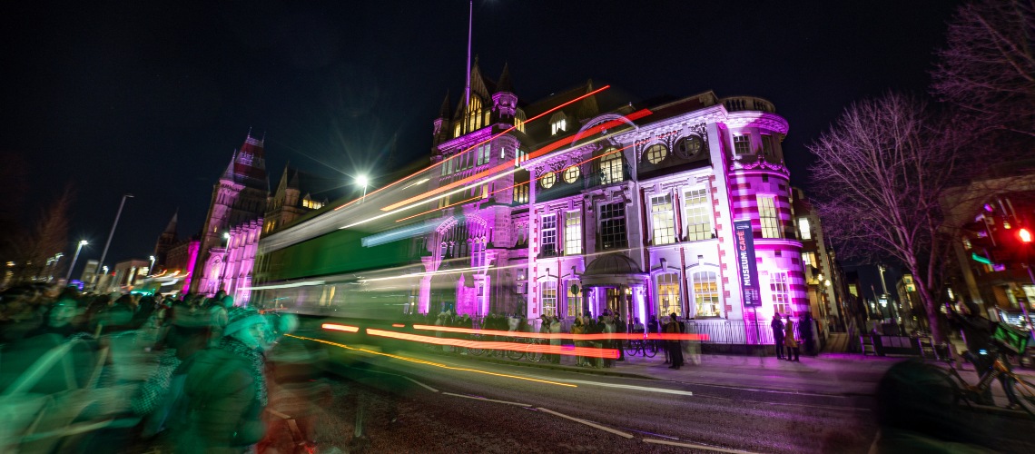 Manchester Museum lit up in purple with a bus going past and a crowd of people in front.
