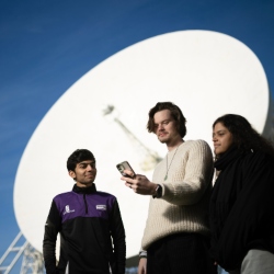 Three students look at a smartphone in front of the Lovell Telescope.