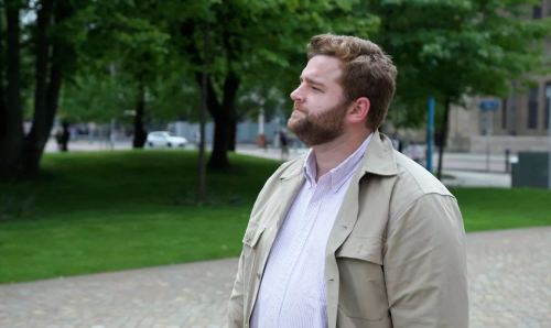 Male Manchester graduate with brown hair and wearing white shirt and brown jacket looks away from camera