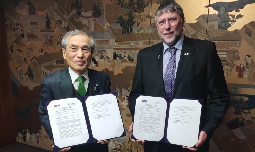 One man from Osaka and one from Manchester stood together holding documents towards the camera.