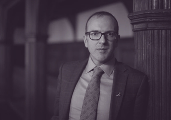 Black-and-white portrait of a man in a suit and glasses, standing beside a wood-panelled column