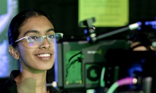 Woman wearing glasses smiling near camera and broadcast equipment with illuminated screens