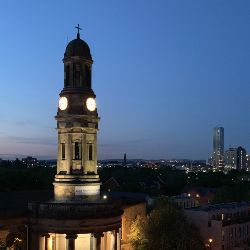 Saint Philips Chapel Street, Salford lit against a dark dusk/night sky, with some of the tall elements of the Manchester skyline in the background.