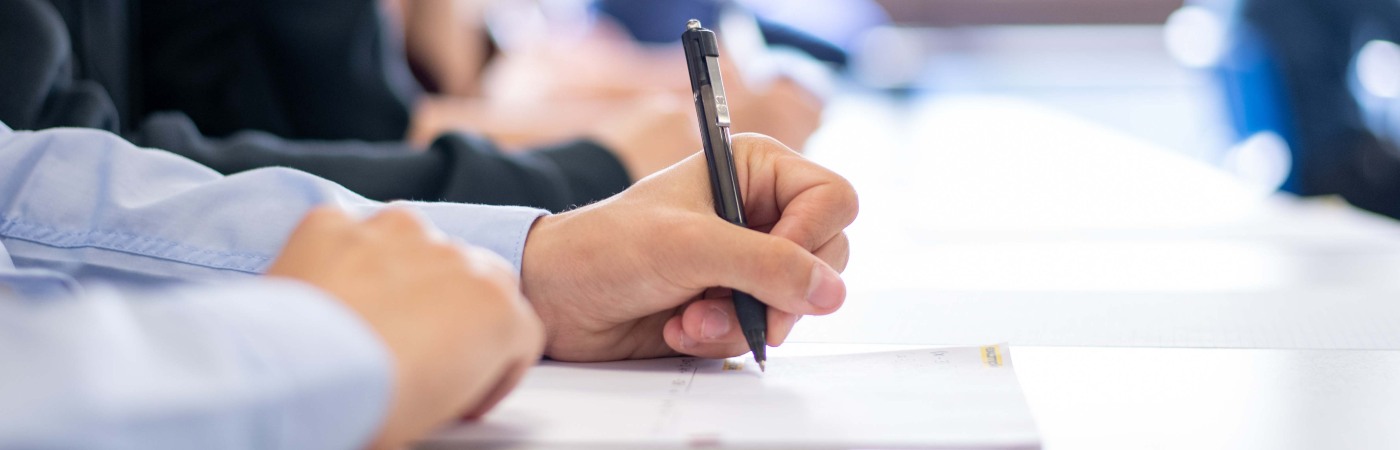 iStock image of student in blue shirt writing on paper with a pen.