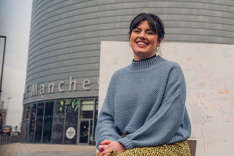 A student sat and smiling in front of the Tin Can building on Oxford Road.