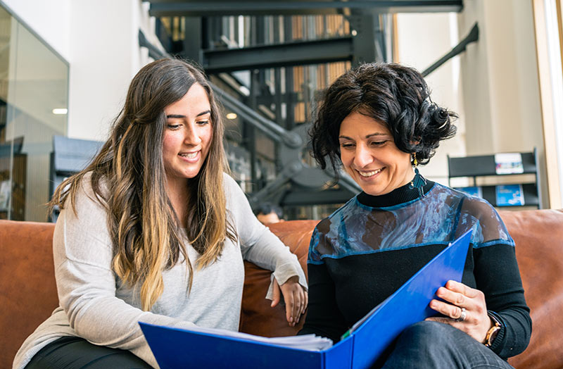 Two women sat on a couch smiling and reading a folder.