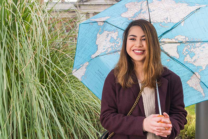 A student smiling and holding an umbrella with a map of the world design.