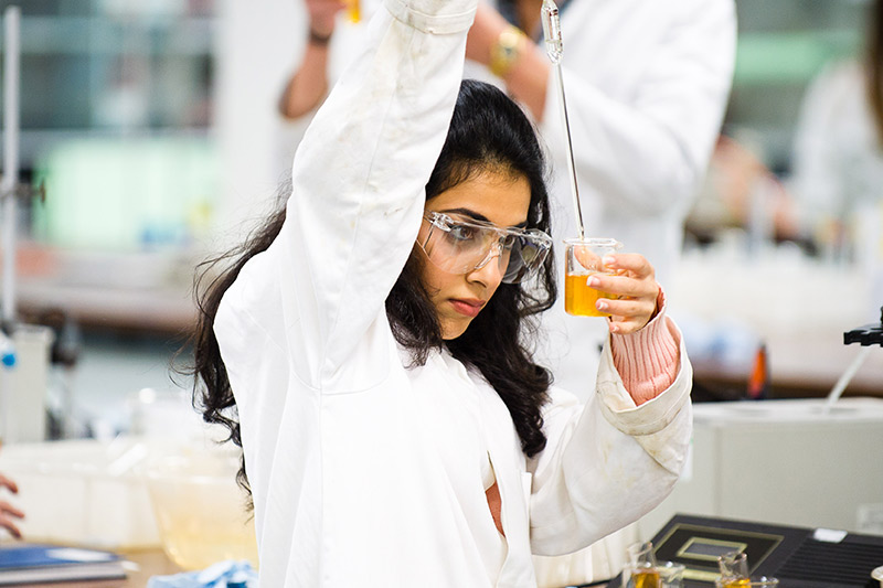 A student in lab coat and protective glasses inspecting liquid in a beaker.