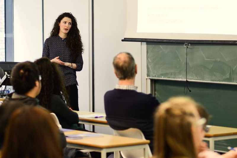 A teacher points to the board while speaking to students in a lecture theatre.
