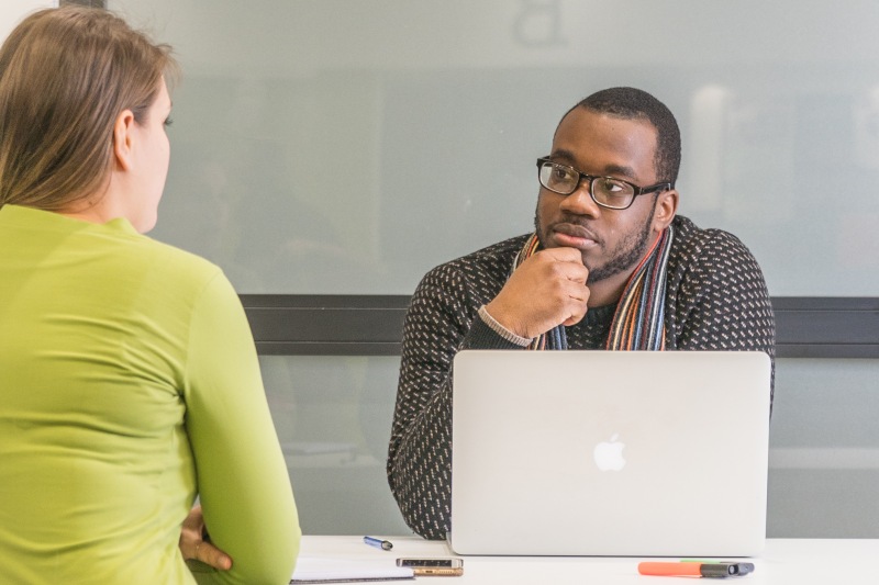 A man with a laptop in conversation with a woman facing him.