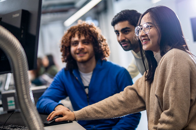 Three students smile as they gather round a desktop computer.