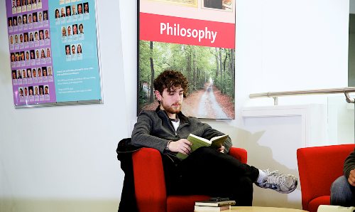 Male philosophy student sat reading a book in front of 'philosophy' sign. 