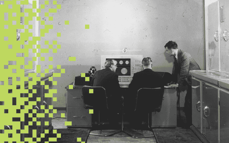 Two men sit at the control panel of the Ferranti MK I, with Alan Turing stood to the right.