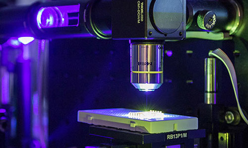 A close-up of the lens of a microscope positioned above a sample plate, backlit by a light.