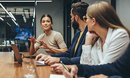 Woman leading a business meeting, speaking to colleagues around a conference table in a modern office.