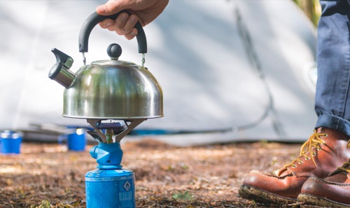 A hand reaches down to pick up a kettle on a gas camping stove.