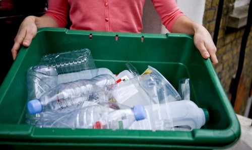 Person carrying green recycling box with assortment of empty plastic bottles.