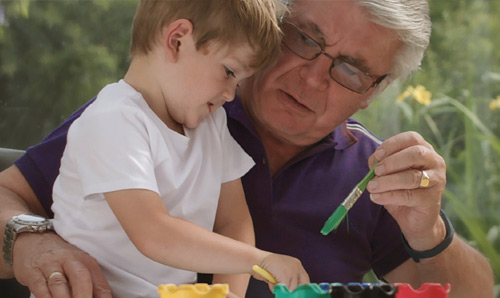 An older man holds a young child while handing him a crayon.