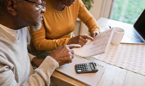 An older Black couple both point at a piece of paper that contains information about bills. There is a laptop and calculator on the table below them.