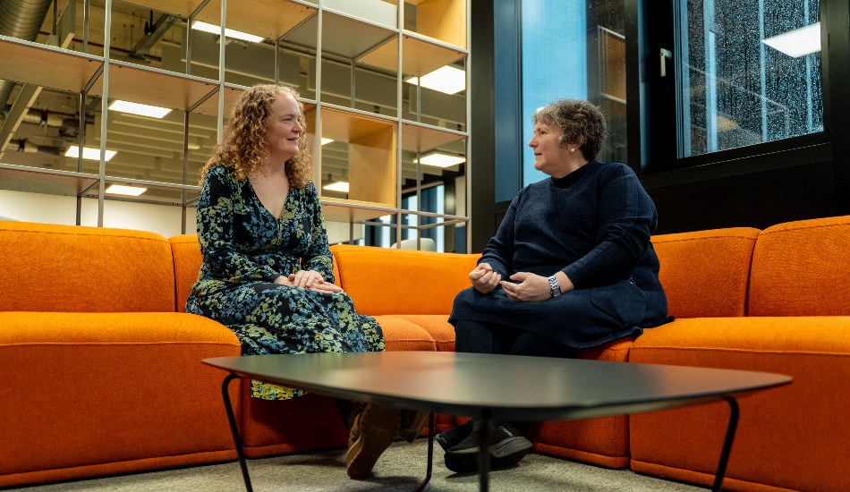 Two people sitting on an orange sofa indoors, talking in a modern lounge area.