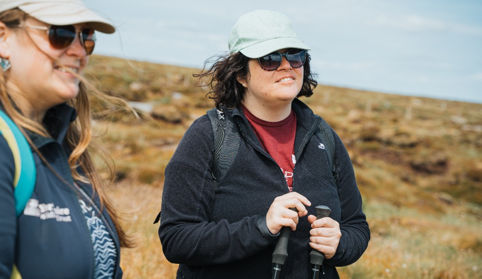 Two people outdoors on moorland, wearing hats and sunglasses, standing with walking poles.