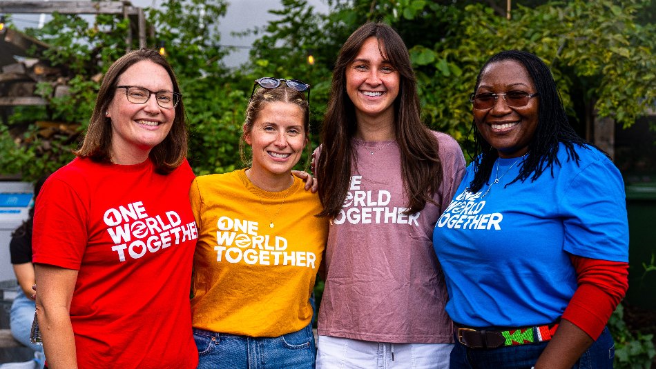 Four people standing together outdoors, smiling, wearing &ldquo;One World Together&rdquo; T-shirts.