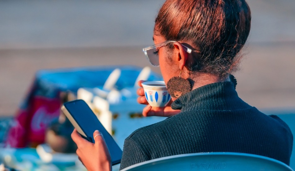 A person sitting outdoors, holding a phone and a cup, viewed from behind.