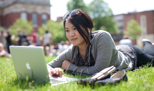 A student lying on a field looking at their laptop.
