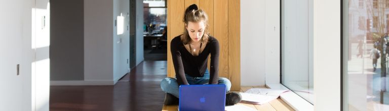 A student sat working on a laptop by a large window.