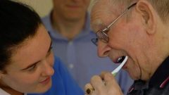 A healthcare professional helping an older man brush his teeth. 