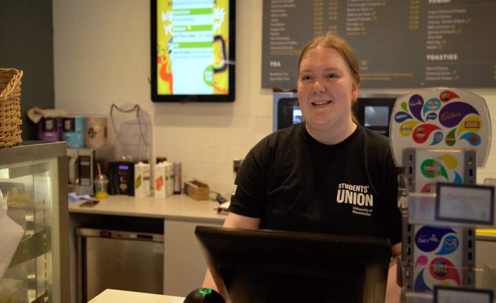 Student working behind a café counter, wearing a black Students’ Union T-shirt, with drinks machines and snack display beside them.