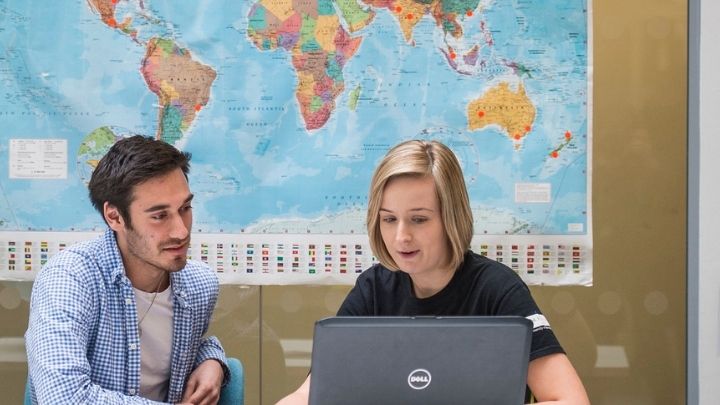 Man and woman sitting in front of laptop with world map in background.