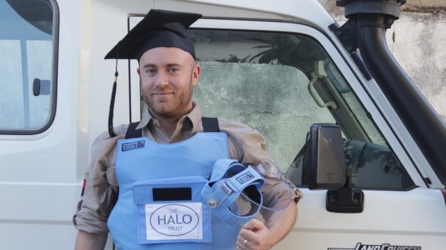 Man stood in front of a HALO Trust branded 4x4 wearing protective equipment and a mortar board.